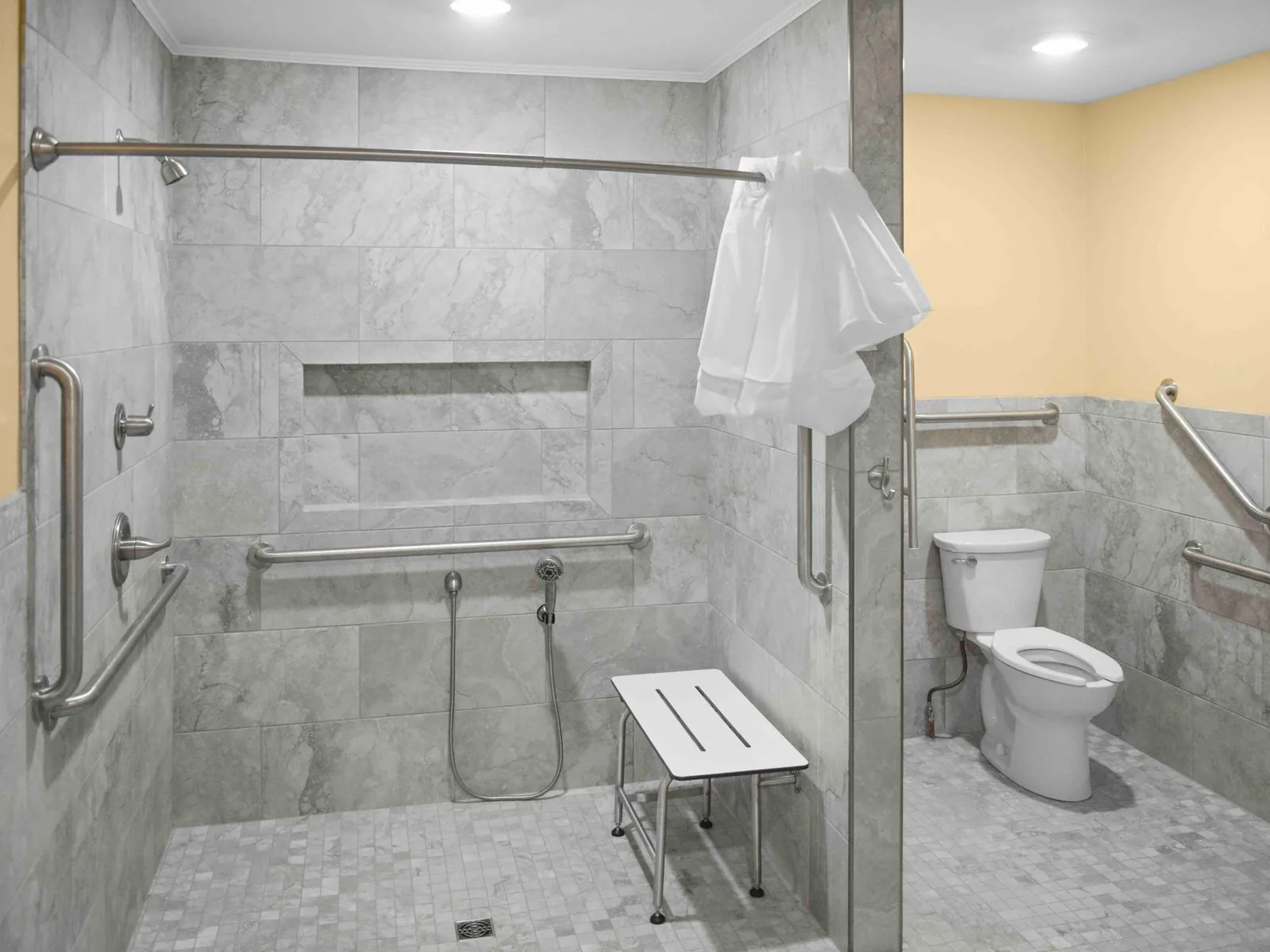 Accessibility remodeling: a curbless tiled roll in shower with grab bars, a handheld shower and a shower bench beside a toilet with grab bars