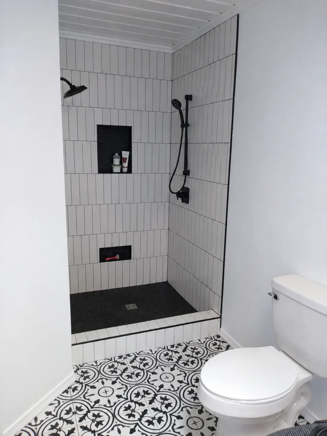 Shower remodeling: a walk in shower with white vertical subway tile, black fixtures, two tiled niches and a patterned tile floor